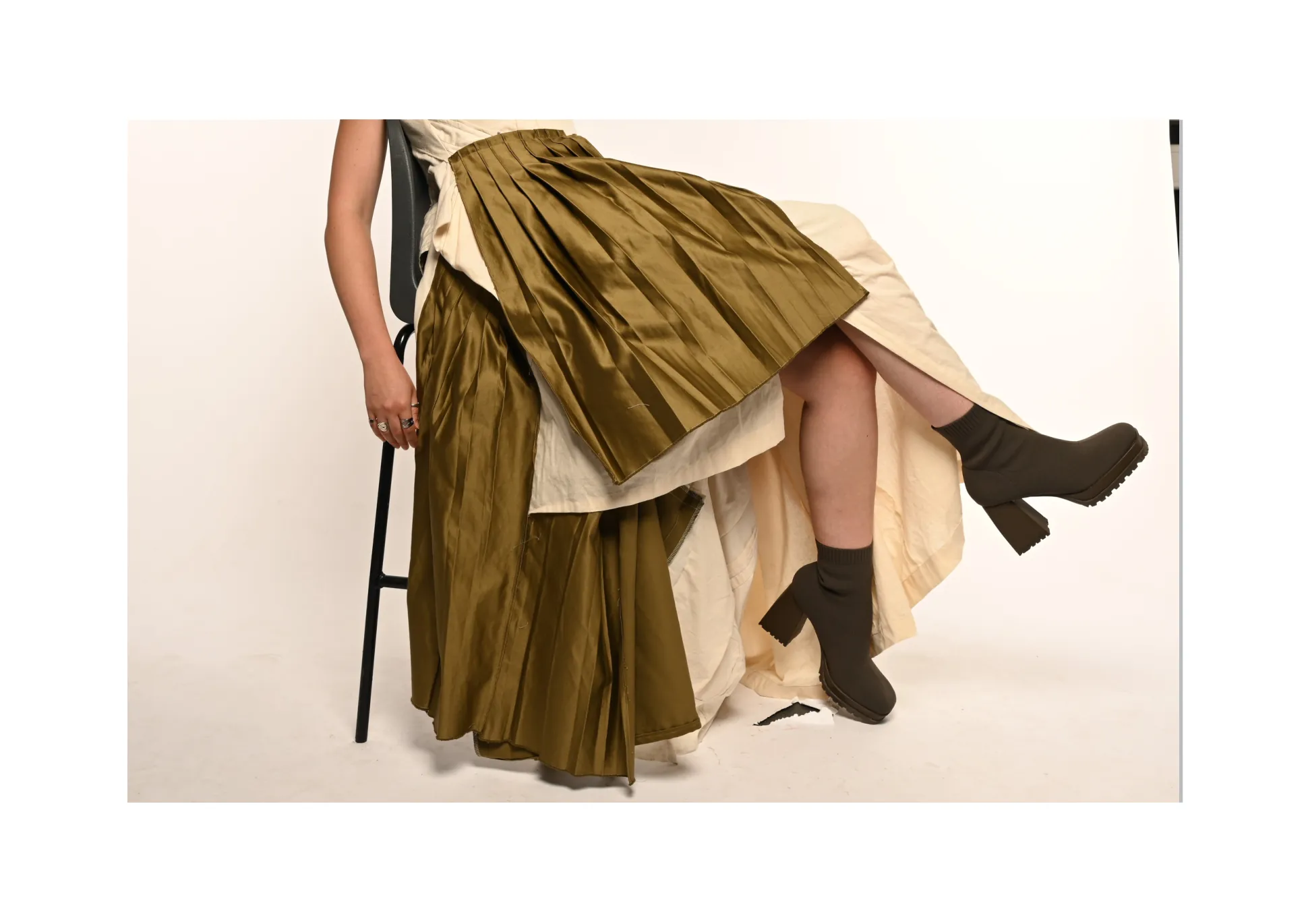 Look-book spread — seated, the olive pleated silk skirt layered over the cream cotton gown, worn with chunky boots.