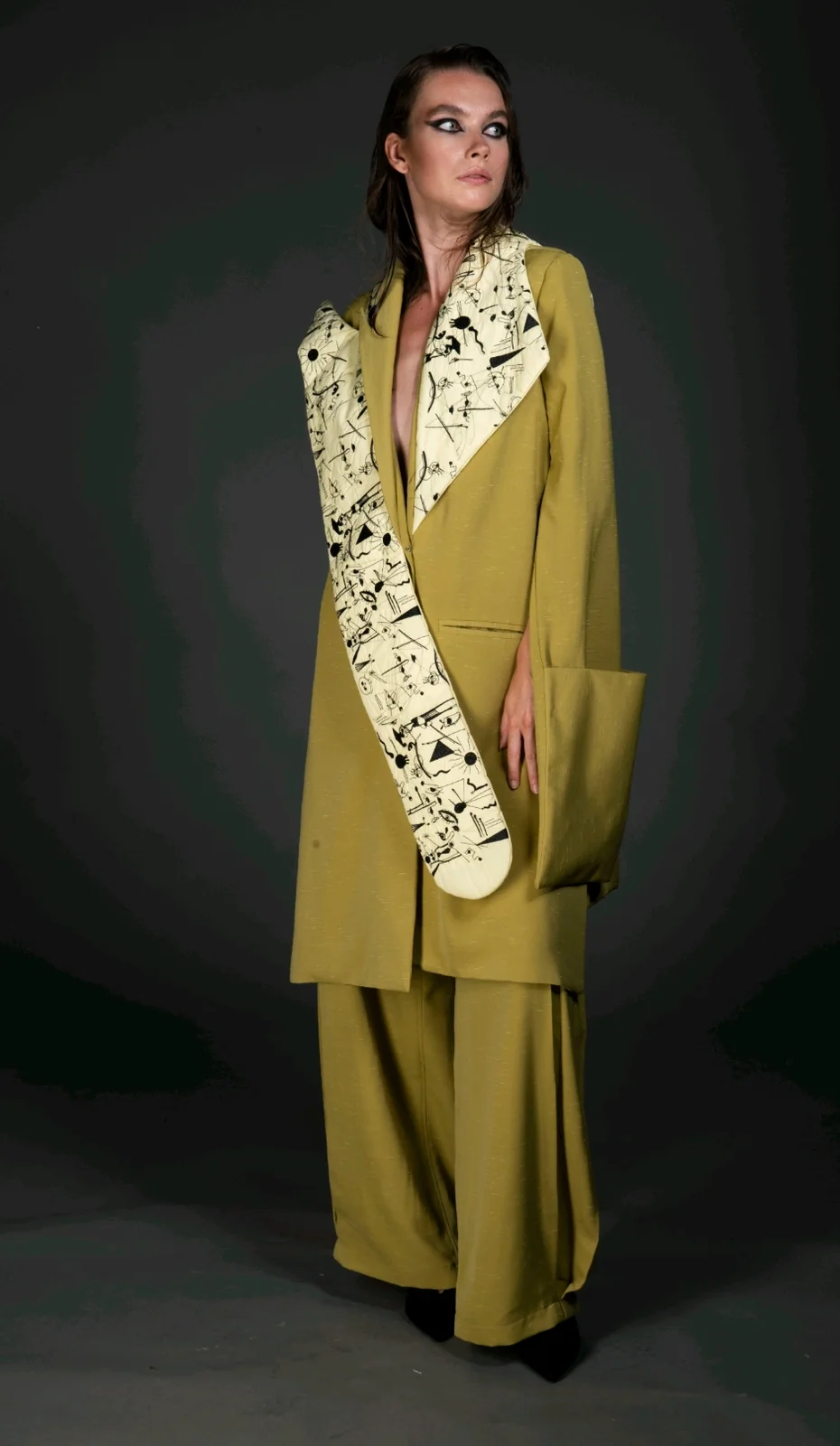 Three-quarter view — the oversized dusted-olive coat and wide trousers, doodle-print panel falling from the shoulder.